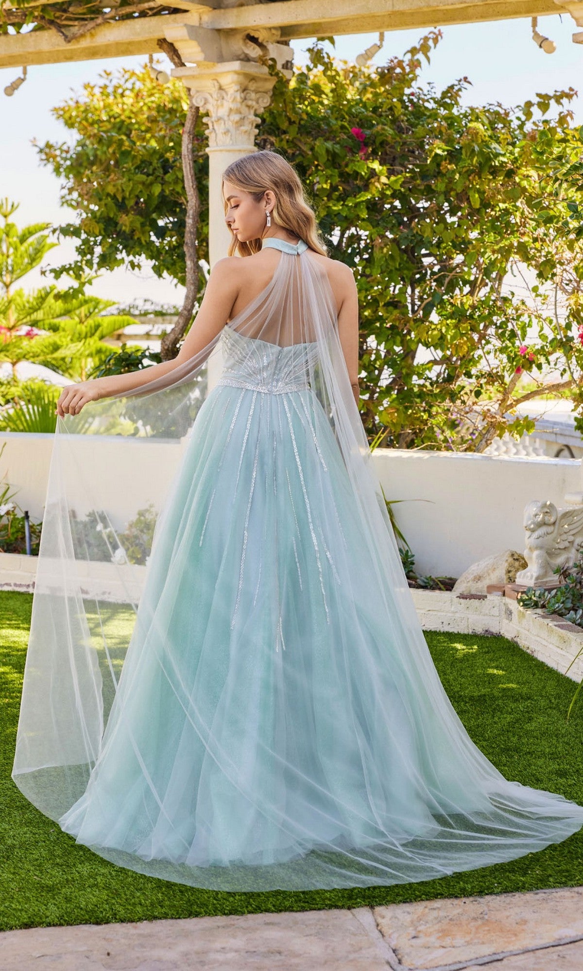 A woman stands outdoors on grass, facing away from the camera, wearing the Juliet JT2620U long prom dress—a light blue, floor-length A-line gown with a sheer tulle overlay and sparkly rhinestone-embellished bodice.